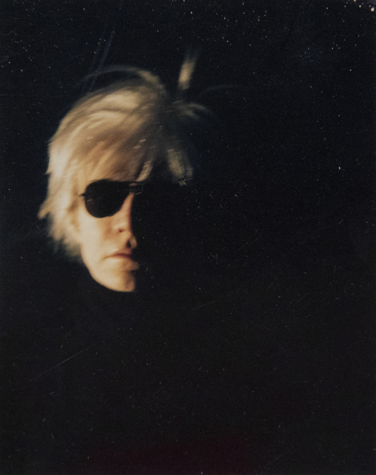 Beginning in 1963, with a silkscreen derived from a photo booth strip, Warhol repeatedly explored his likeness, culminating with the iconic "Fright Wig" image. Essential to his depiction of celebrities and self-representation, the Polaroid photograph played a crucial role in his work and our perceptions of his massive contribution to post-war art in America. The two images presented, dark and spooky, are beautifully crafted, well-staged portraits. Enveloped in a moody ambiance that eviscerates his body, these self-portraits depict Andy clad in this iconic wig and dark aviator sunglasses, set against a backdrop so deeply shadowed that his head seems to float in a void of darkness. Warhol loved role-playing, and here it is in spades!